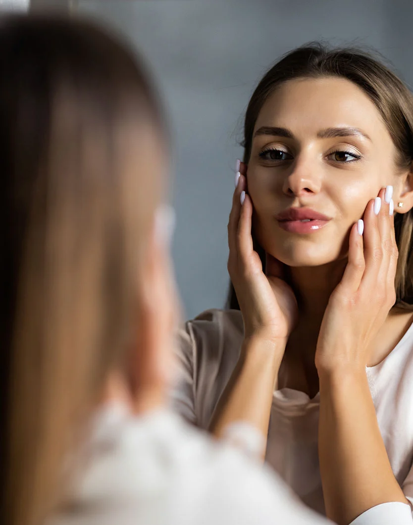 Close-up of a woman gazing at her reflection. She has long brown hair, light skin, and a gentle expression, with hands resting on her cheeks and neck. She wears a light beige top, blurred in the background. The muted gray backdrop keeps the focus on her face and hands in the reflection. - Botox and Wrinkle Relaxers Recovery in Las Vegas, NV