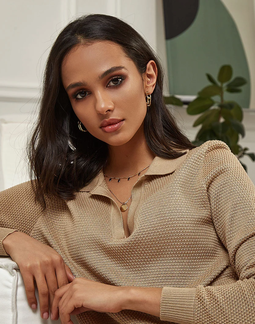 A woman with long, dark brown hair and subtle makeup wears a nude-beige knitted polo shirt with a V-neck and front zip. She has a gold-toned necklace and chain earrings. The background features a neutral off-white wall with a pale green abstract design and plants. Soft lighting highlights her face and upper body. - Dermal Fillers Recovery in Las Vegas, NV